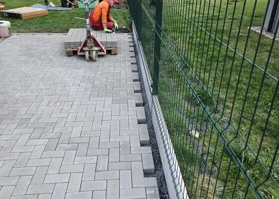Položená zámková dlažba podél plotu, v pozadí paleta s dlažbou