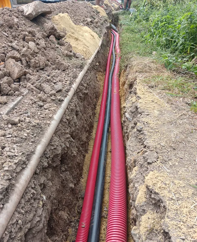 Dvě červené chráničky a černý kabel na pískovém loži ve výkopu