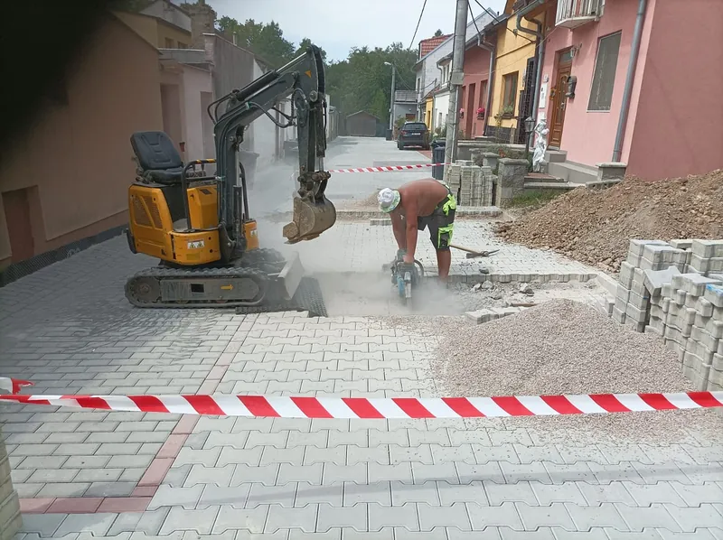 Malé rypadlo v ulici a pracovník řeže dlažbu, vedle palety zámkové dlažby